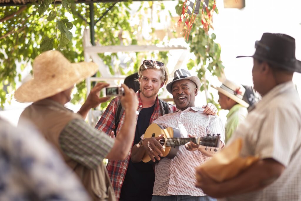Market trader taking photograph of tourist posing with market trader playing ukulele