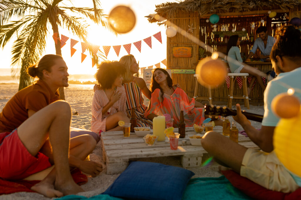 A multi-ethnic group of people enjoying their time at a beach with their friends during a sunset, sitting on sand, playing guitar and smiling